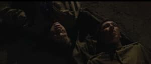Two soldiers lying on the ground at night, with serious expressions, in a military setting.