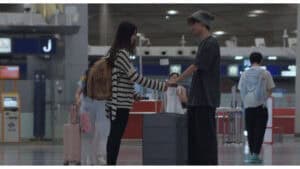 Airport greeting, young couple with luggage at check-in, casual travel attire, busy terminal scene.