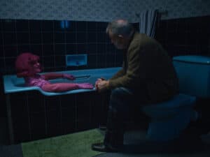 Whimsical woman in pink vintage outfit with glove and wig, talking to man on toilet in dimly lit bathroom.