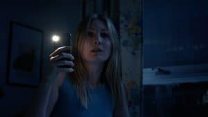 Asian woman taking selfie with mobile phone in dark room, illuminated by phone flash, expressing surprise or shock.