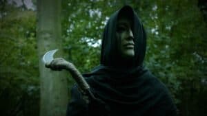 Eerie figure with mask and scythe in dense forest for Halloween or horror theme.